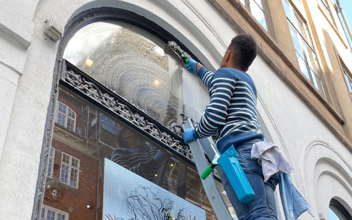 Mand i blå stribet sweatshirt og blå handsker pudser stort frontvindue fra stige