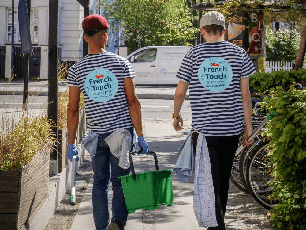 To vinduespudsere med stribede t-shirts og French Touch logo, begge bærer rengøringsartikler